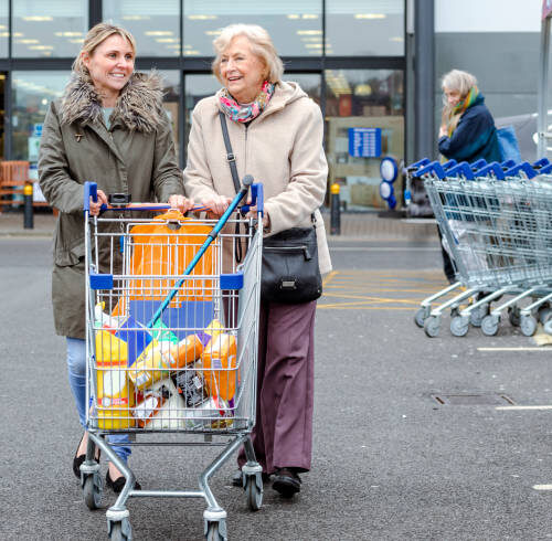A mature woman helps a senior woman from the shops with her shopping trolley