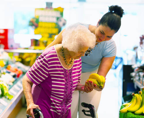 Young hispanic caregiver supporting senior woman, 93 years, with buying banans in a shop