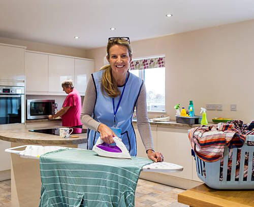 Care worker making a home visit. Female carer is ironing in the kitchen to help an elderly woman. The elderly woman is standing at the kitchen bench behind but not in focus.